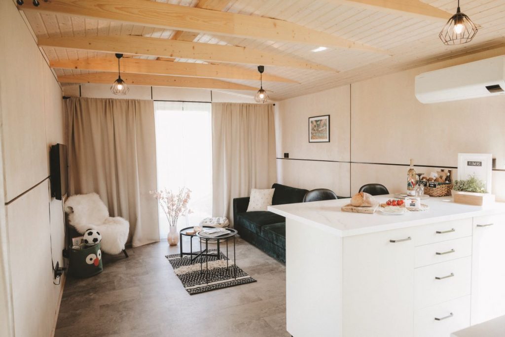 Obývačka za bielym kuchynským ostrovom so smaragdovou sedačkou, konferenčnými stolíkmi a televízorom na stene.