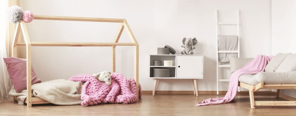 Detská drevená posteľ s konštrukciou v tvare strechy domu s ružovým vankúšom a dekou na drevenej podlahe. Vedľa nízka biela skrinka na drevených nôžkach, biely rebrík a svetlosivá sedačka na drevenej konštrukcii.