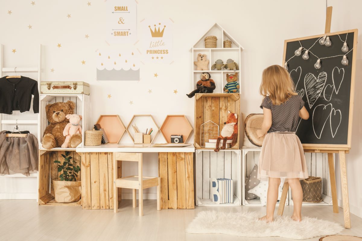Kreatívny kútik v detskej izbe s bielo-dreveným nábytkom. Drevené prepravky slúžia ako úložný priestor i nohy stola pre malé dievčatko.