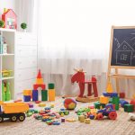 Detská izba s bielou policou, bielou komodou, čiernou tabuľou a farebnými hračkami, rozsypanými na zemi.