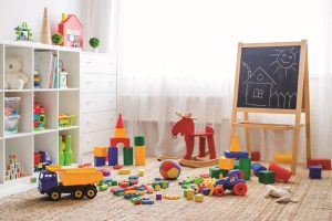 Detská izba s bielou policou, bielou komodou, čiernou tabuľou a farebnými hračkami, rozsypanými na zemi.