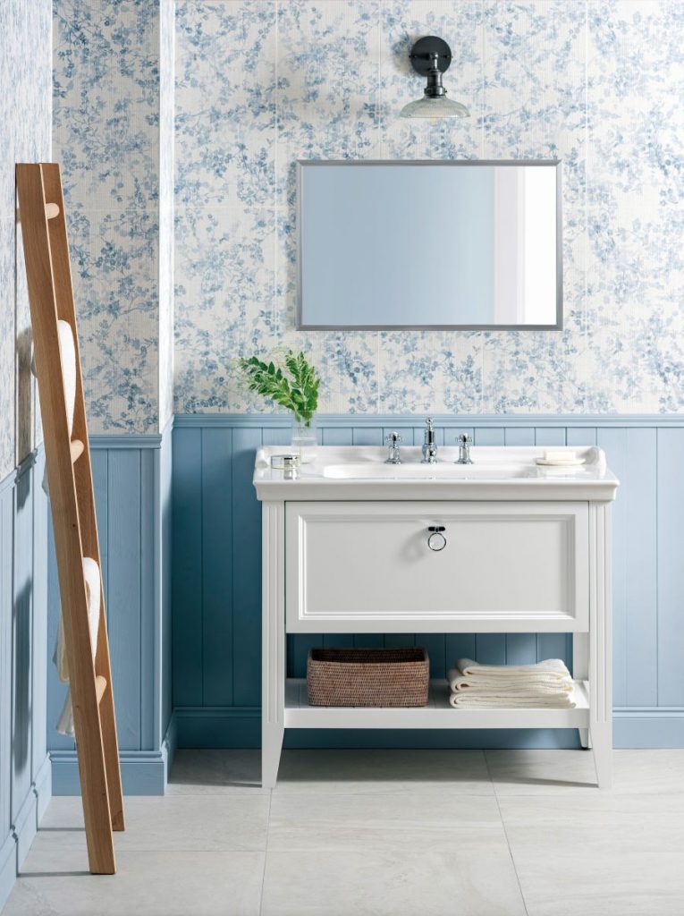 Kúpeľňa s modrým zvislým obkladom do tretiny výšky steny, vyššie kvetinový vzor na stenách. Uprostred samostatne stojace umývadlo a nad ním zrkadlo.
