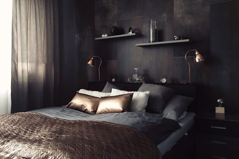 Manželská posteľ v sivočiernej miestnosti s bronzovými ozdobnými vankúšmi a bronzovou dekou.