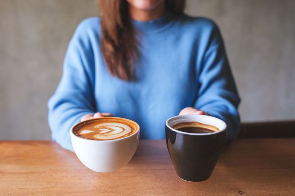 Žena v modrom pulóvri drží pred sebou dve kávy, jedna je čierna klasika. Tá druhá má latte art.