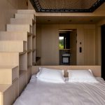 Interiér chatky z dreva s manželskou posteľou, schodmi s úložným priestorom a otvorenými dvere do kúpeľne.