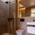 Kúpeľňa v chate so sprchovacím kútom s drevenou podlahou, toaletou, umývadlom a obdĺžnikovým zrkadlom s podsvietením po obvode.
