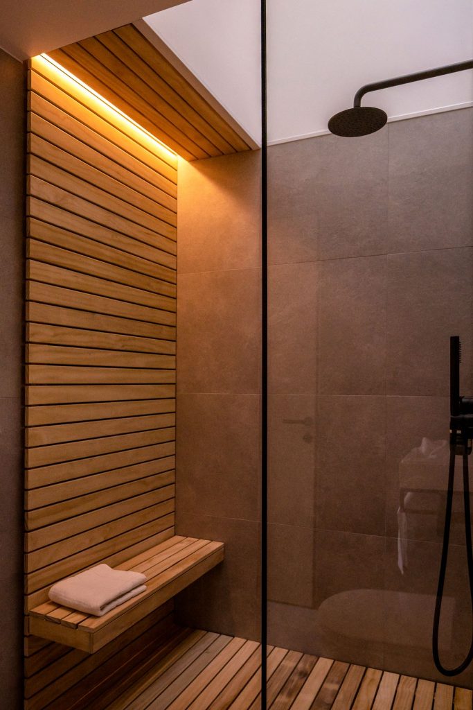 Sprchovací kút so sklom s čiernym rámom, čiernou hlavicou a malou drevenou lavičkou na protiľahlej stene.
