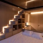 Osvetlený interiér chatky z dreva s manželskou posteľou, malou kuchynkou a schodmi s úložným priestorom, vedúce k sieti v mezaníne.