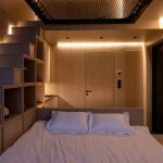 Osvetlený interiér chatky z dreva s manželskou posteľou a schodmi s úložným priestorom, vedúce k sieti v mezaníne.