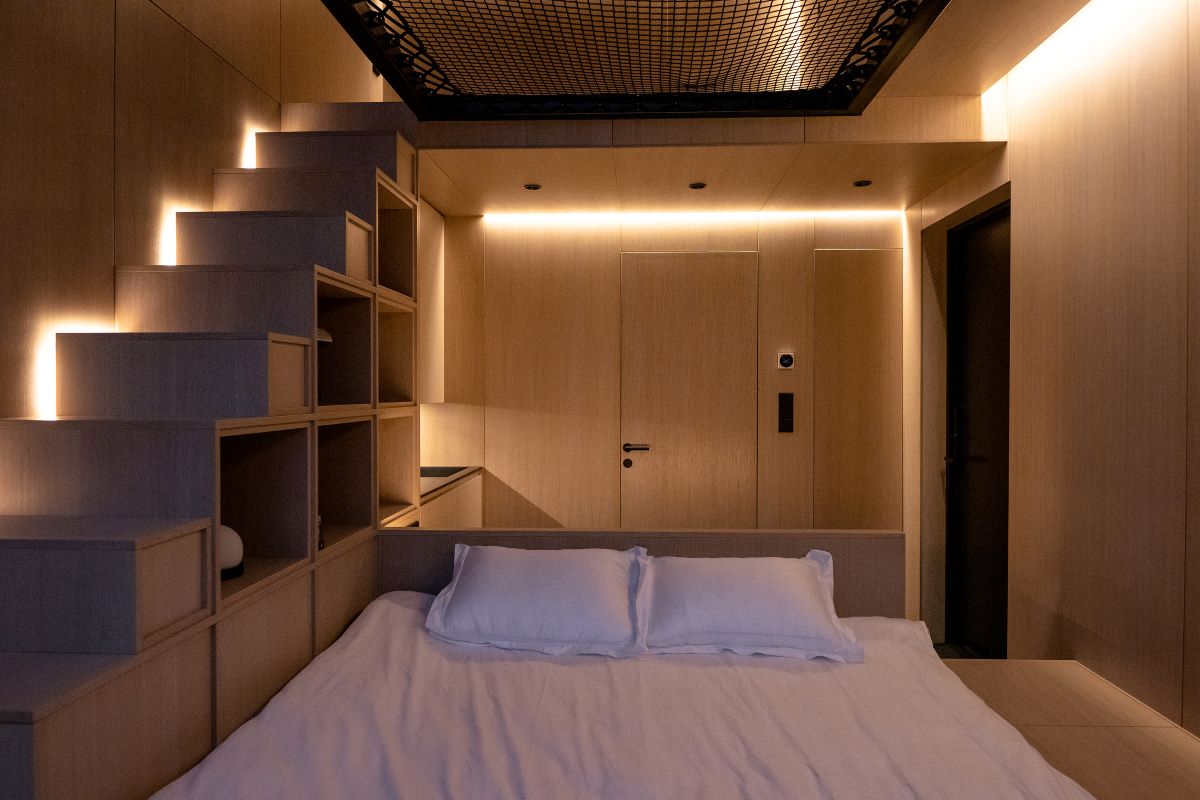 Osvetlený interiér chatky z dreva s manželskou posteľou a schodmi s úložným priestorom, vedúce k sieti v mezaníne.