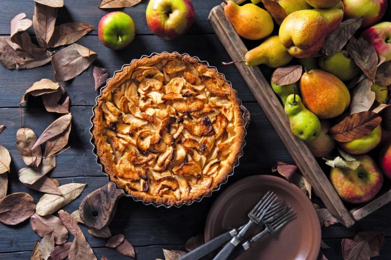Jablkový pie vo vrúbkovanej forme na stole, obložený lístím, jablkami a hruškami.