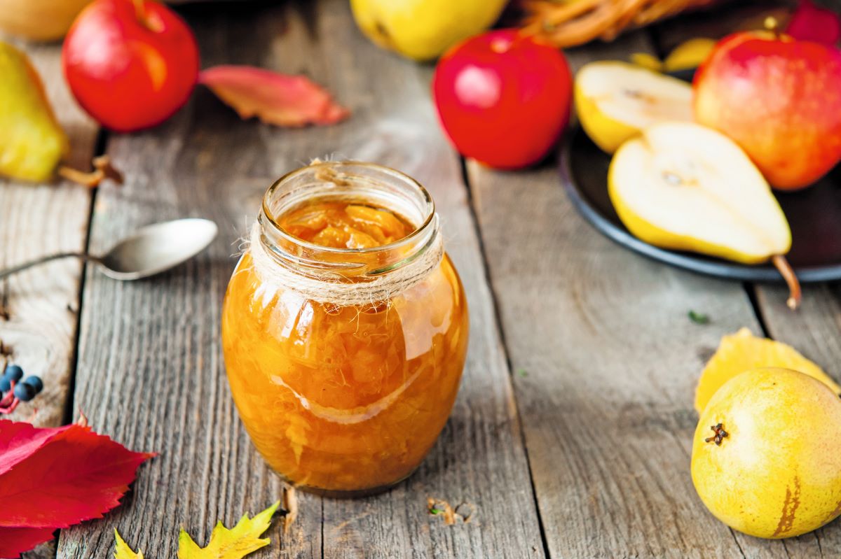 Jablkovo-hruškový džem v pohári na drevenom stole, obklopený lístím a celými jablkami a hruškami.