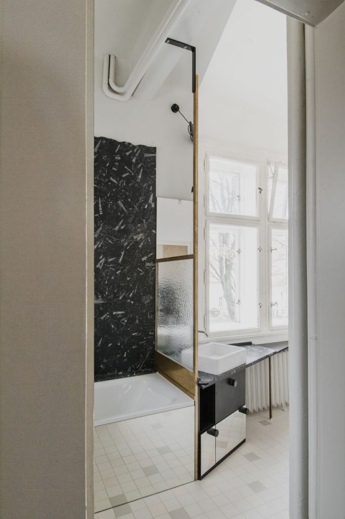 Kúpeľňa v byte, ladená do čierno-biela, s vaňou so zrkadlovým obkladom a asymetricky umiestneným umývadlom pod oknom.