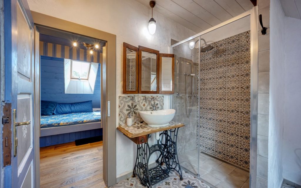 Kúpeľňa na statku s umývadlom na starom stolíku, trojdielnym zrkadlom s dreveným rámom a sprchovacím kútom so vzorovanými kachličkami.