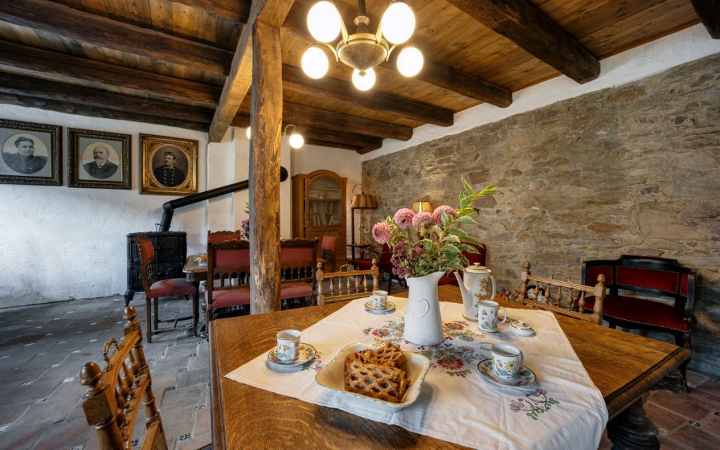 Kuchyňa s pohľadovými trámami, jedným štvorcovým jedálenským stolom s drevenými stoličkami a ďalším obdĺžnikovým stolom s čalúnenými stoličkami.