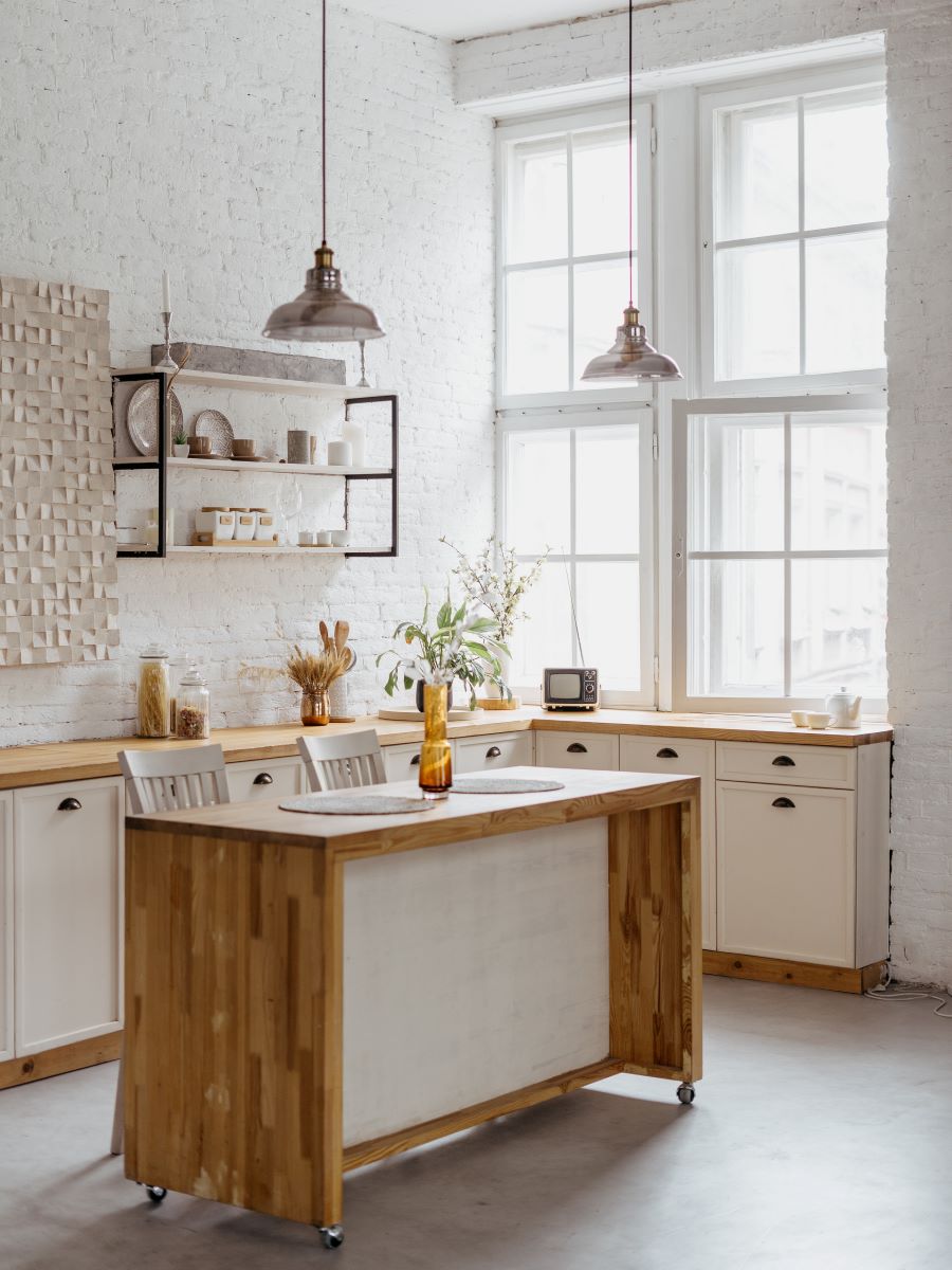 Vidiecky zariadená kuchyňa v bielo-drevenom prevedení s lampami zavesenými nad ostrovčekom, slúžiacim aj ako stôl.