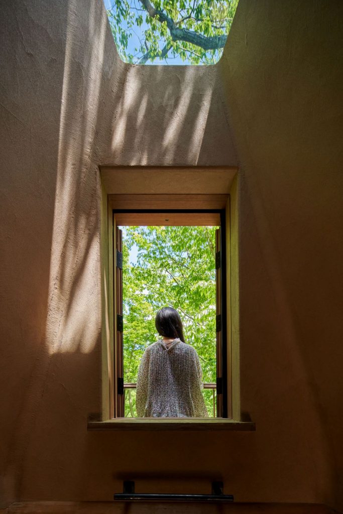 Žena stojí chrbtom k oknu na balkóniku na streche a díva sa na les.