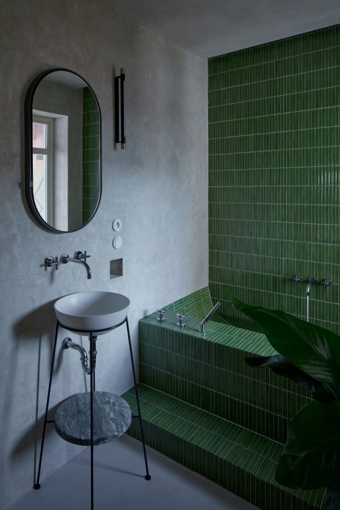 Jedna z kúpeľní v byte s vaňou obloženou zeleným obkladom, oválnym zrkadlom a atypickým umývadlom.