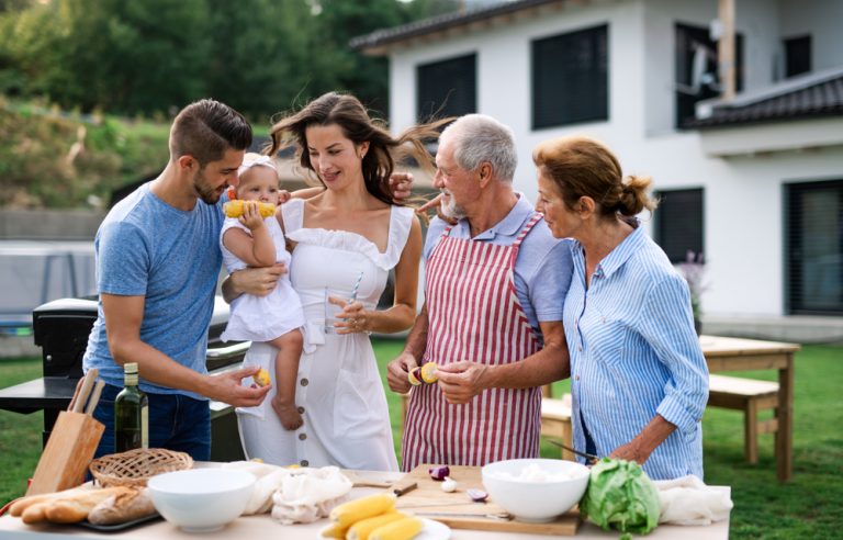 Fotografia trojgeneračnej rodiny so starými rodičmi, rodičmi a malým dievčatkom na rukách matky pri stole s jedlom v exteriéri.
