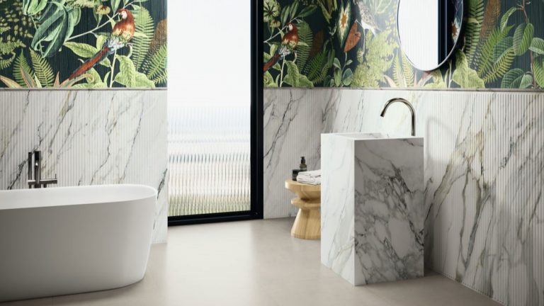 Umývadlo v tvare mramorového kvádra s jamkou v strede a samostatne stojaca biela vaňa v kúpeľni s farebnými stenami s tropickým vzorom.
