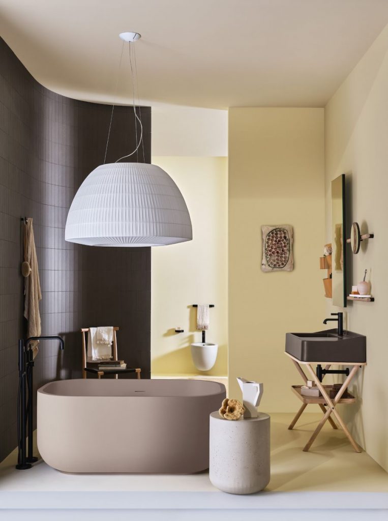 Samostatne stojaca vysoká vaňa, smerujúca kolmo na zaoblenú stenu žlto-čiernej kúpeľne. Doplnená je o čierne hranaté umývadlo, valcový stolík a stropné svietidlo.