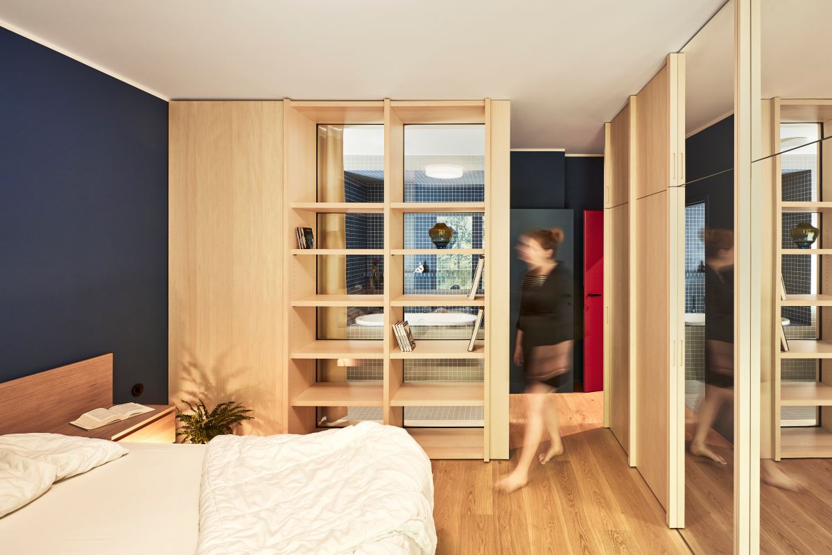 Spálňa s prevažne dreveným nábytkom i policou s vertikálnym presklením a tmavomodrou stenou za čelom postele.
