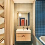 Kúpeľňa pri spálni s modrou a šedomodrou mozaikou na stenách i podlahe, bielym umývadlom, drevenou skrinkou a zrkadlom.