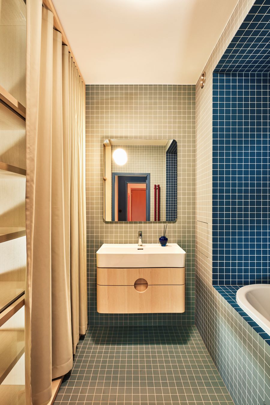 Kúpeľňa pri spálni s modrou a šedomodrou mozaikou na stenách i podlahe, bielym umývadlom, drevenou skrinkou a zrkadlom.