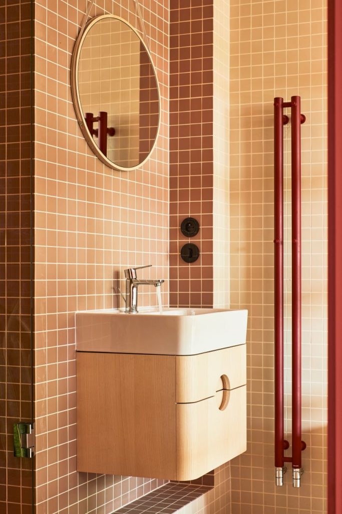 Kúpeľňa s hnedou štvorcovou mozaikou, bielym umývadlom na stene, drevenou skrinkou a okrúhlym zrkadlom.