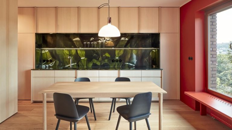 Kuchyňa s obkladom so svetlého dreva na podlahe i stenách, svetlou drevenou linkou i jedálenským stolom, čiernymi stoličkami a jednou červenou stenou s lavičkou pri okne.