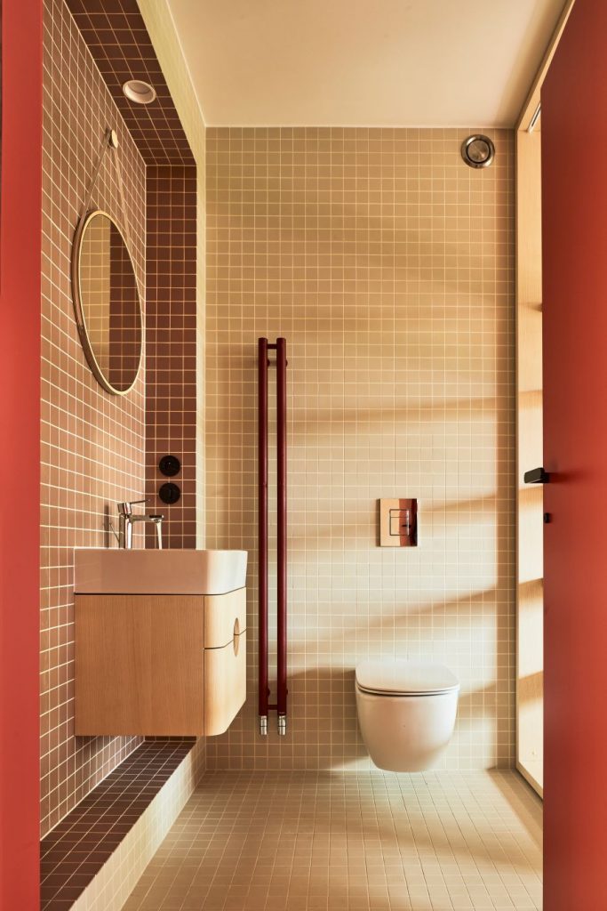 Kúpeľňa s hnedou štvorcovou mozaikou, bielou toaletou i umývadlom na stene, pod ktorým je drevená skrinka. Do kúpeľne vedú červené dvere.