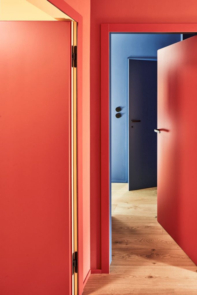 Chodba s drevenou podlahou a červenými i modrými stenami s rovnako farebne zladenými dverami.