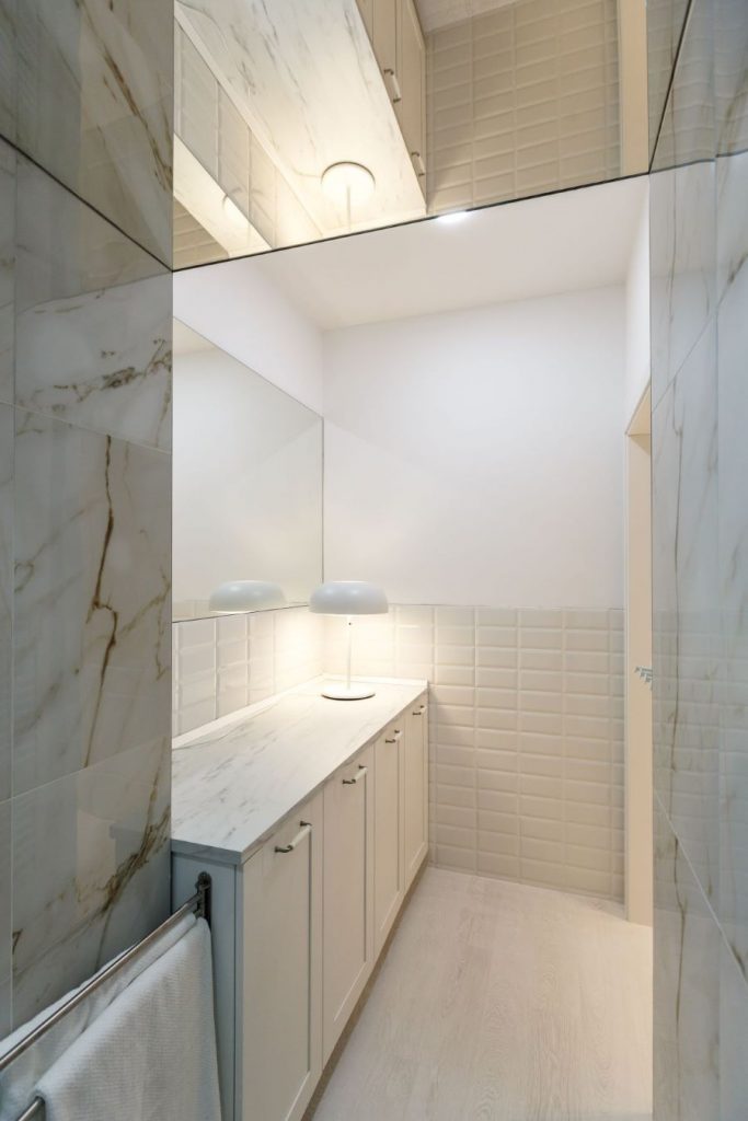 Kúpeľňa v byte s veľkým bezrámovým zrkadlom na stene a bielymi skrinkami, slúžiacimi aj ako polica na odkladanie predmetov.