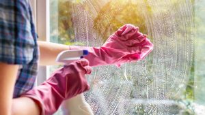 Žena umýva okno čistiacim prostriedkom a handrou.