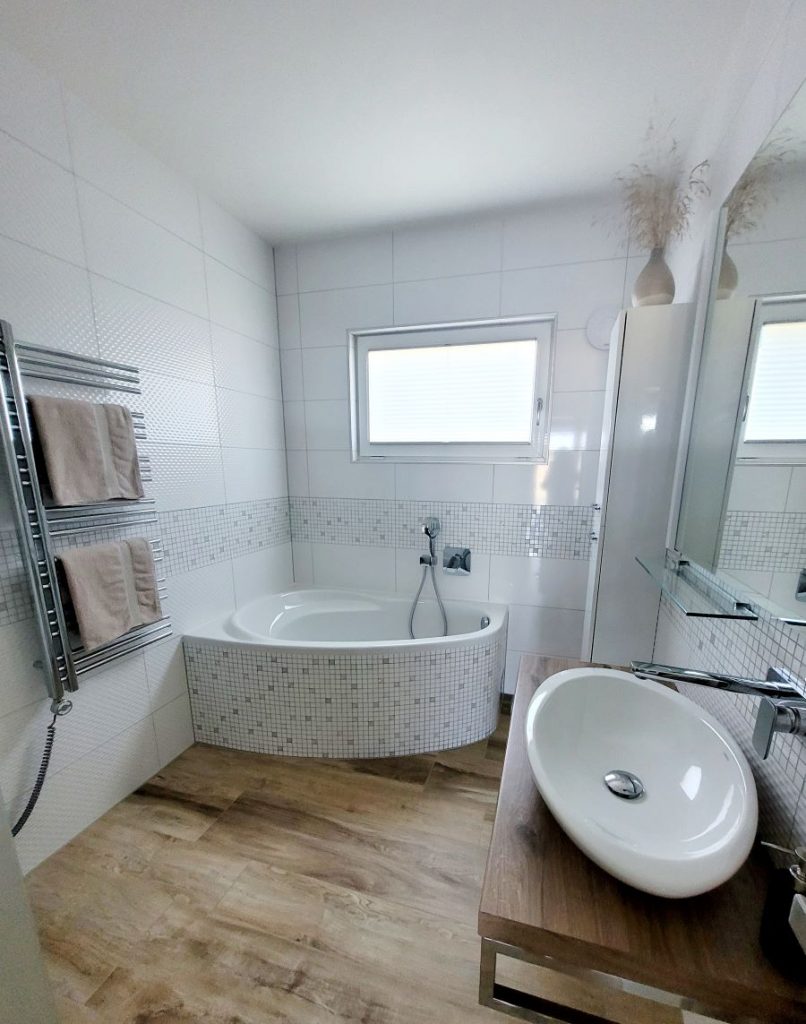 Pohľad do kúpeľne s drevenou podlahou a mozaikovým obkladom na rohovej vani.