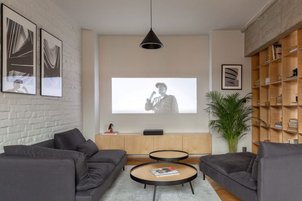 Moderne zariadená obývačka so sivými sedačkami, stolíkmi a priemietaním na stenu.