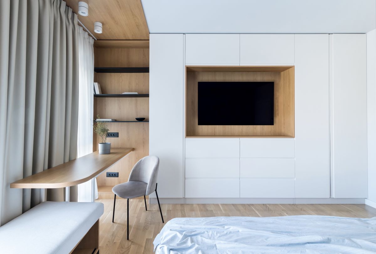 Mnimalisticky zariadená hosťovská izba v neutrálnych odtieňoch s televízorom a úložným priestorom.