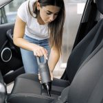 Žena s ručným vysávačom čistí sedačku auta.