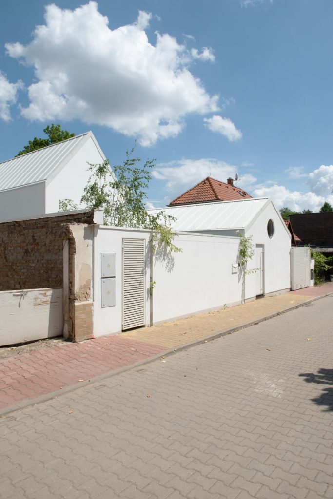Moderná biela budova so sedlovou strechou a bielym murovaným plotom.