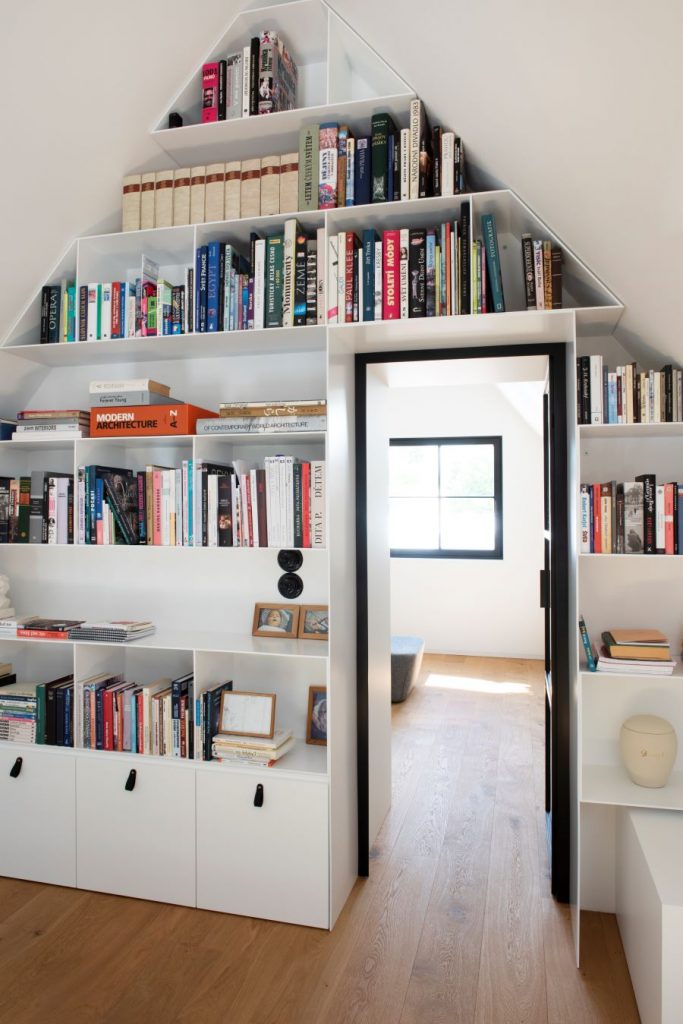 Zostava otvorených políc pod zošikmenou strechou v okolí dverí s knihami.