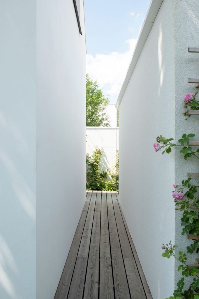 Pohľad na úzky drevený chodník medzi dvoma hmotami bieleho domu.