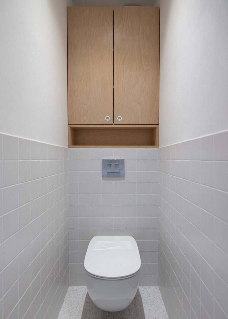 Minimalistické WC s drevenou skrinkou a bielymi obkladačkami.