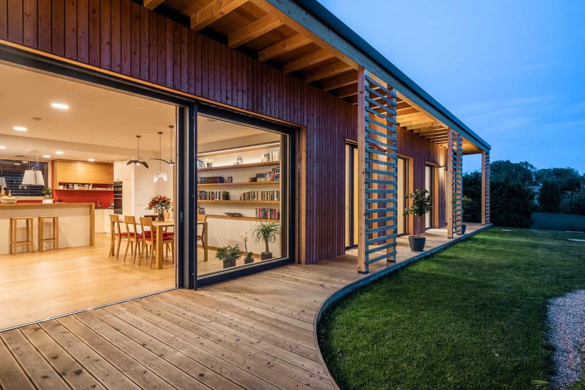 Exteriér moderného dreveného domu so sklenenými dverami a veľkými oknami, večerné osvetlenie terasy a záhrady.
