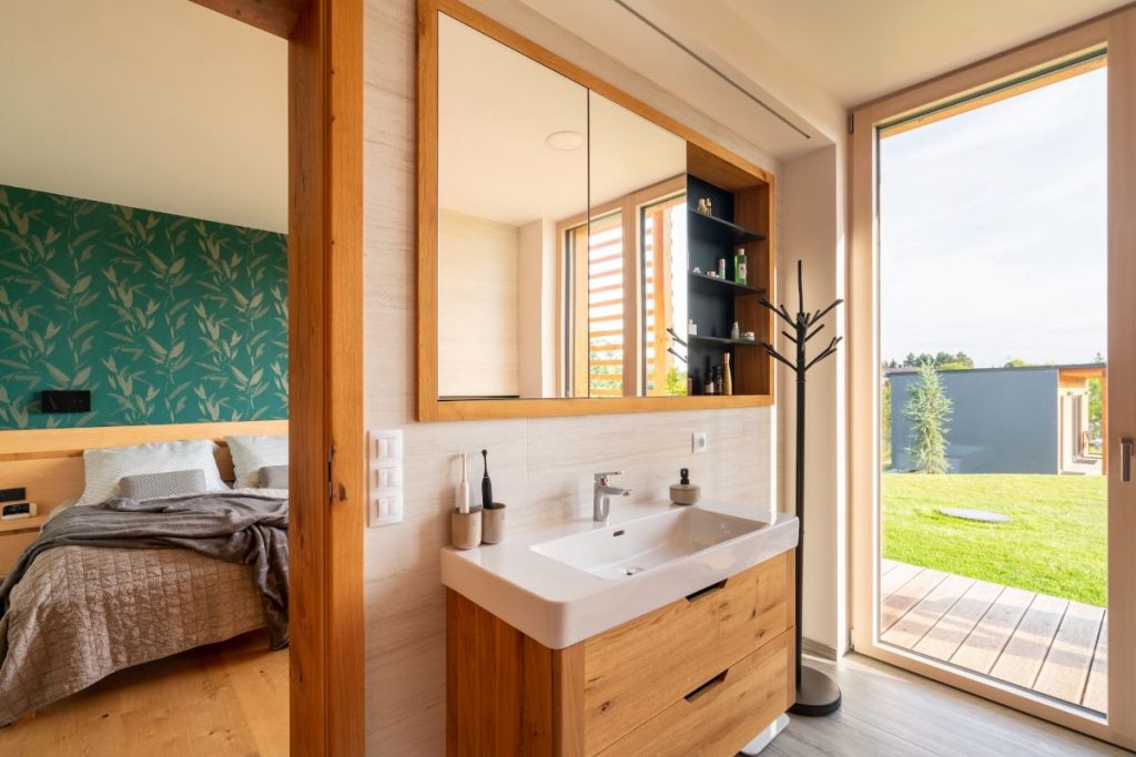 Moderná kúpeľňa s umývadlom a drevenou skrinkou, prepojená so spálňou a so vstupom do záhrady.