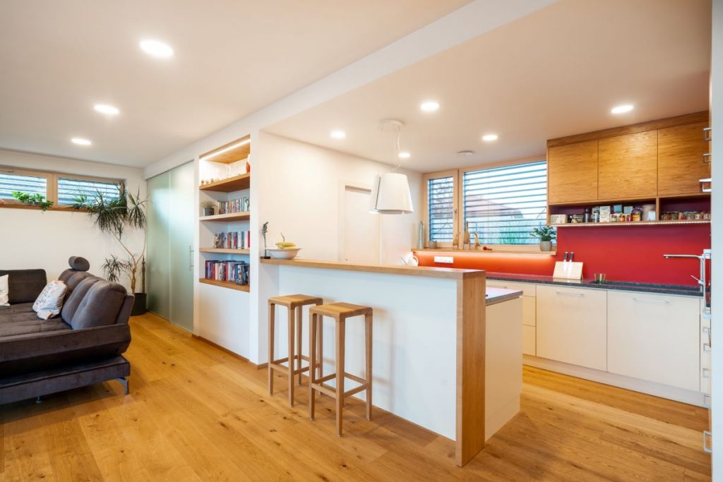 Kuchyňa spojená s obývacou izbou s bielo-drevenou linkou, barovým pultom s dvoma stoličkami a zabudovanou policou na knihy.