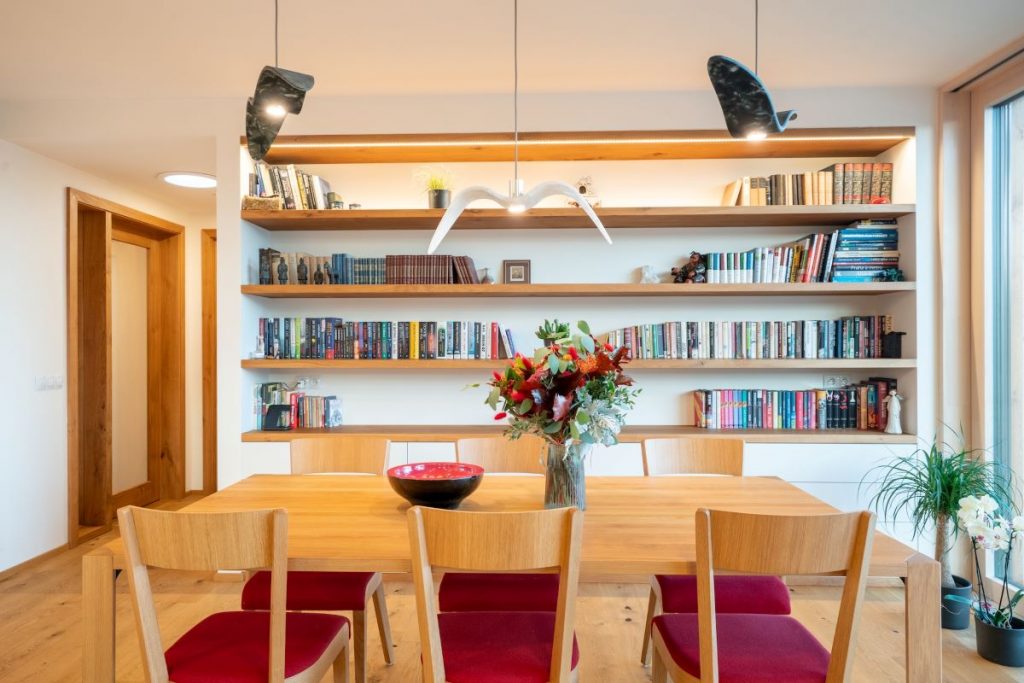 Jedáleň s dreveným stolom a stoličkami s červeným čalúnením, v pozadí otvorené police s knihami.