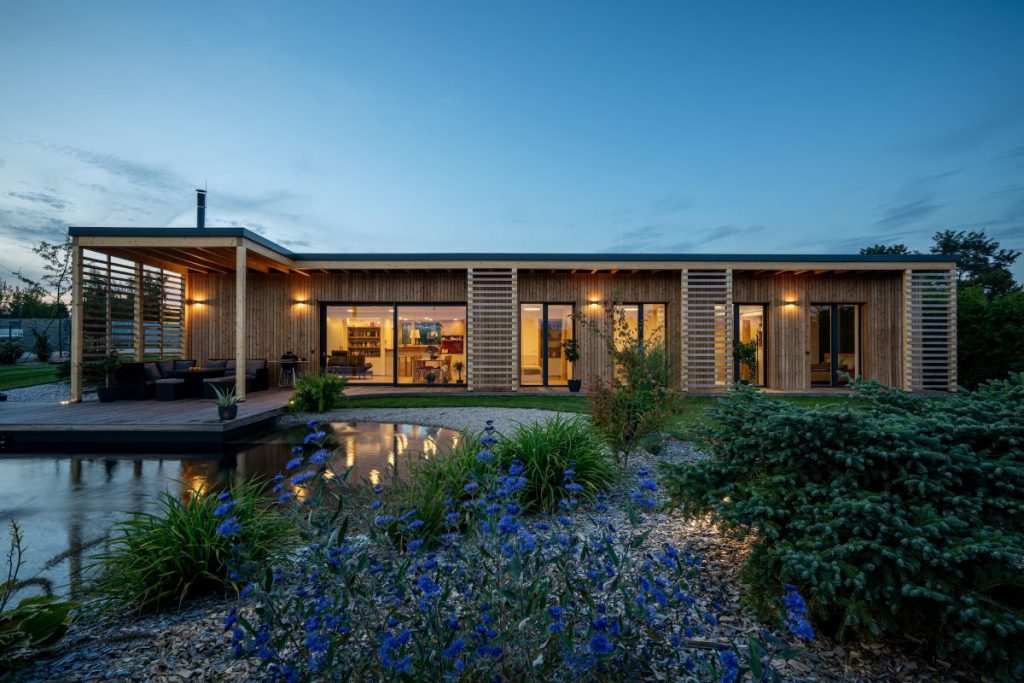 Moderný drevený dom za súmraku so sklenenými dverami a oknami, terasou a záhradou s jazierkom a záhonmi.