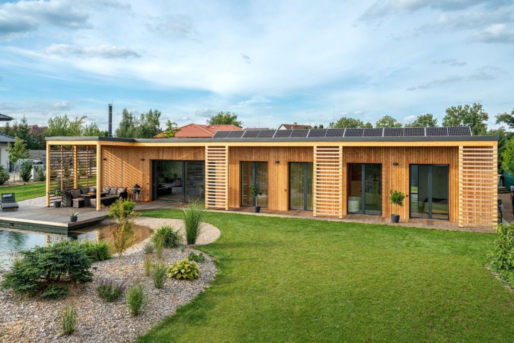 Exteriér dreveného domu s veľkými oknami a fotovoltickými panelmi na streche, v popredí záhrada a vodná plocha.