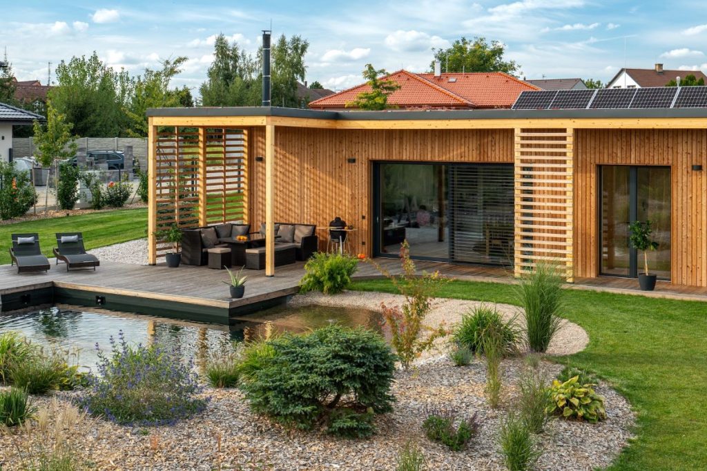 Terasa moderného dreveného domu s nábytkom a ležadlami pri záhrade s vodnou plochou.