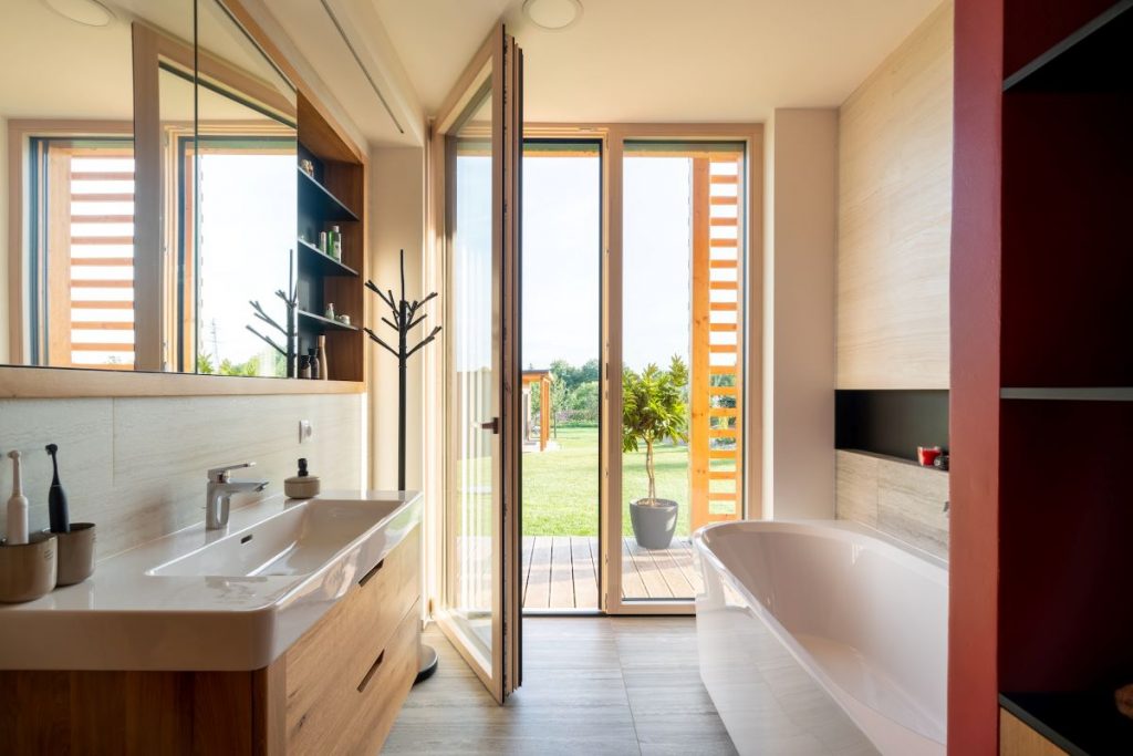 Moderná kúpeľňa s vaňou a umývadlom, dreveným nábytkom a vstupom do záhrady cez sklenené dvere.